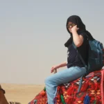 A tourist wearing a black headscarf and sunglasses riding a camel in the Egyptian desert near the Pyramids of Giza, with a colorful saddle and a blue backpack